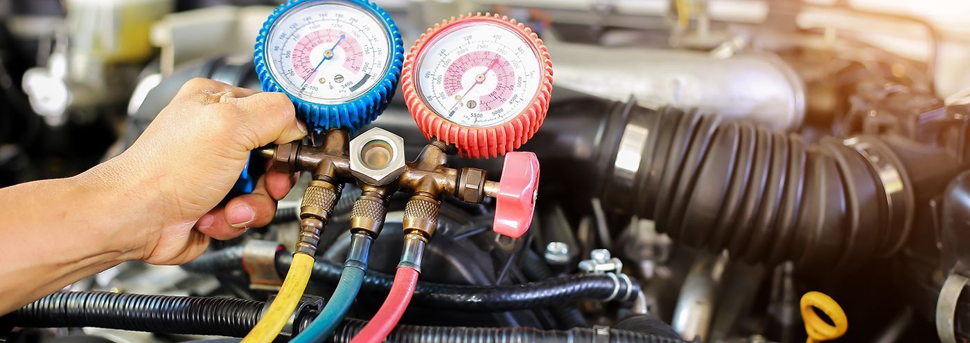 A person's hand connects gauges to an automobile engine.  Blue and red gauges with hoses attached are in the foreground.