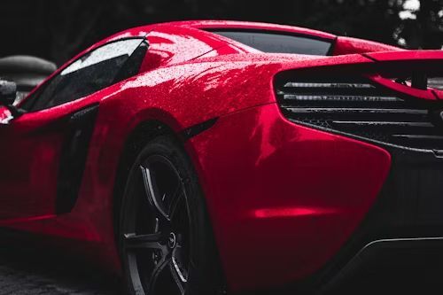 Red sports car, rain droplets on the shiny body, parked outdoors.