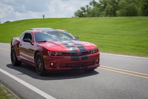 Red Chevrolet Camaro sports car racing on a paved road next to a grassy hill.