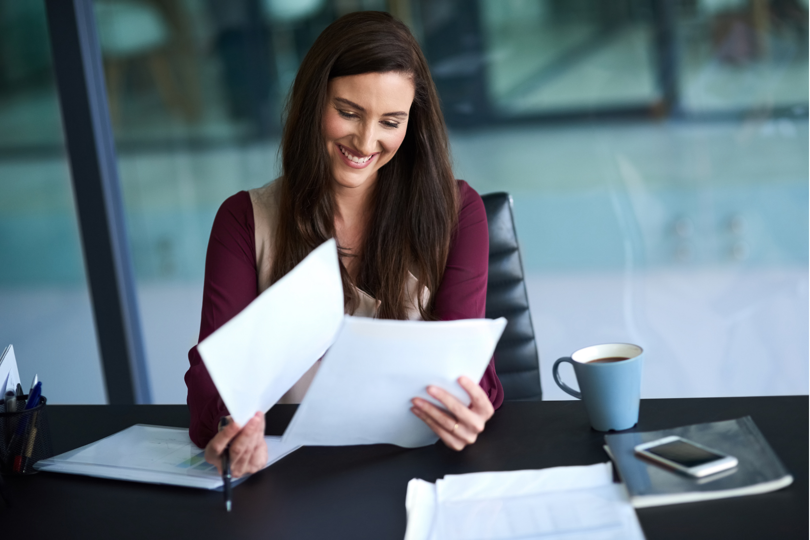 A smiling person sits at a desk, reviewing a document with a coffee mug and phone nearby.