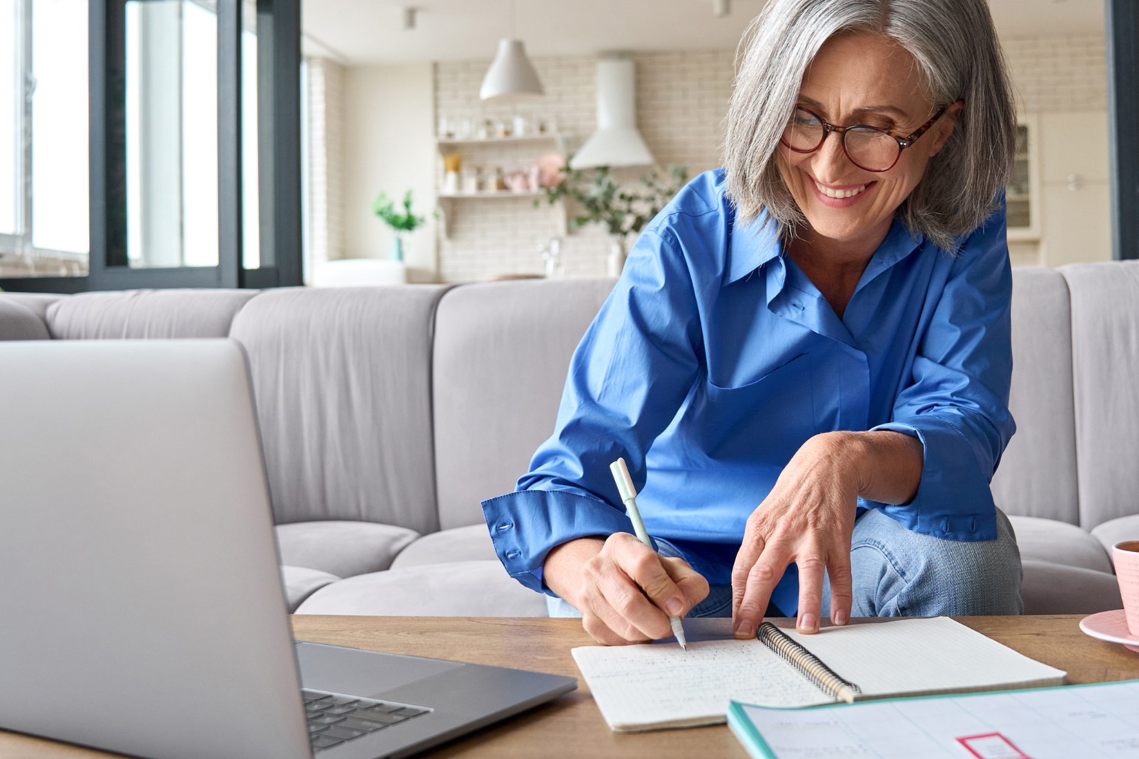 A Virtual Assistant is sitting at a table with a laptop and writing in a notebook.