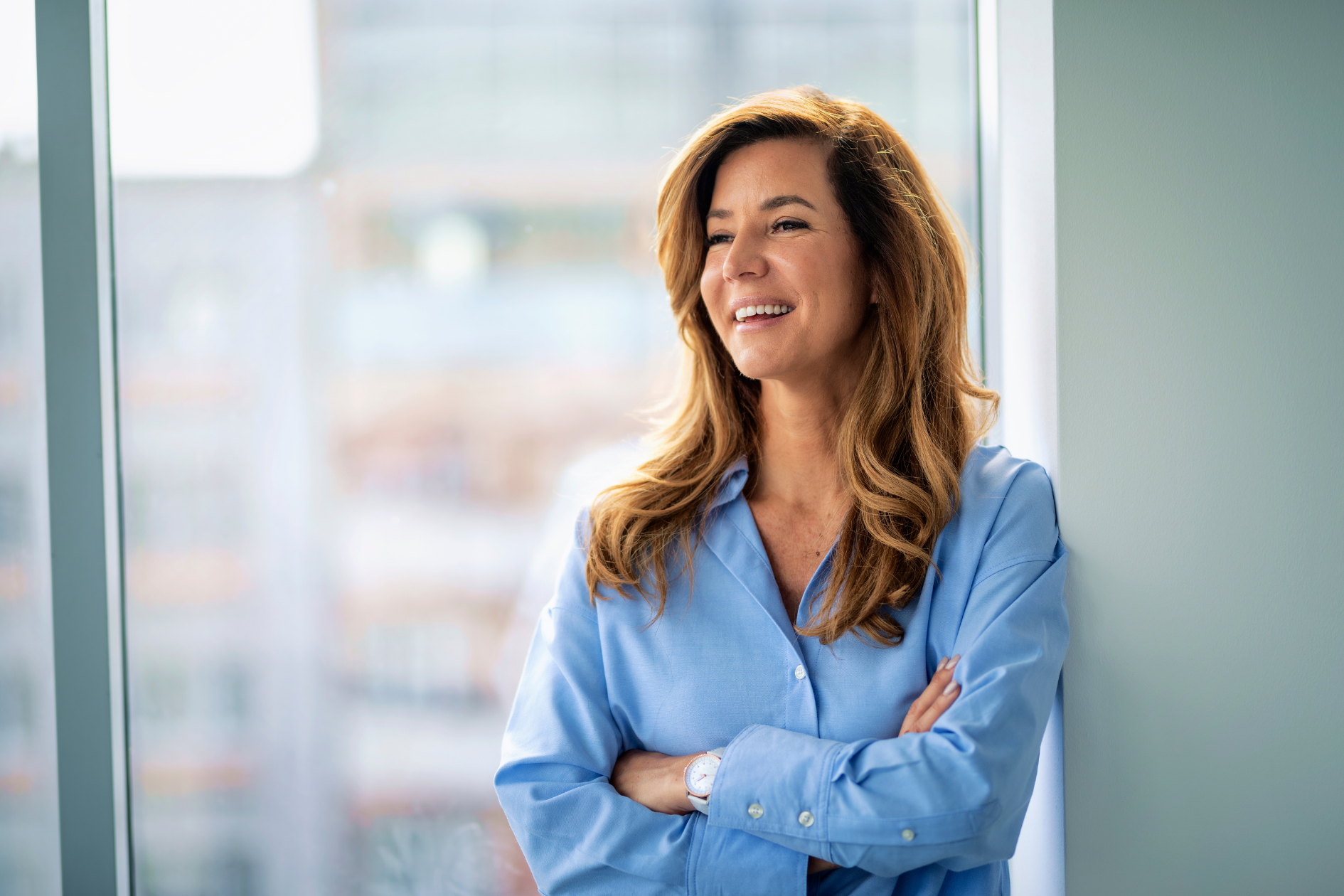 A smiling person with long hair, wearing a light blue button-down shirt, stands with arms crossed by a window.