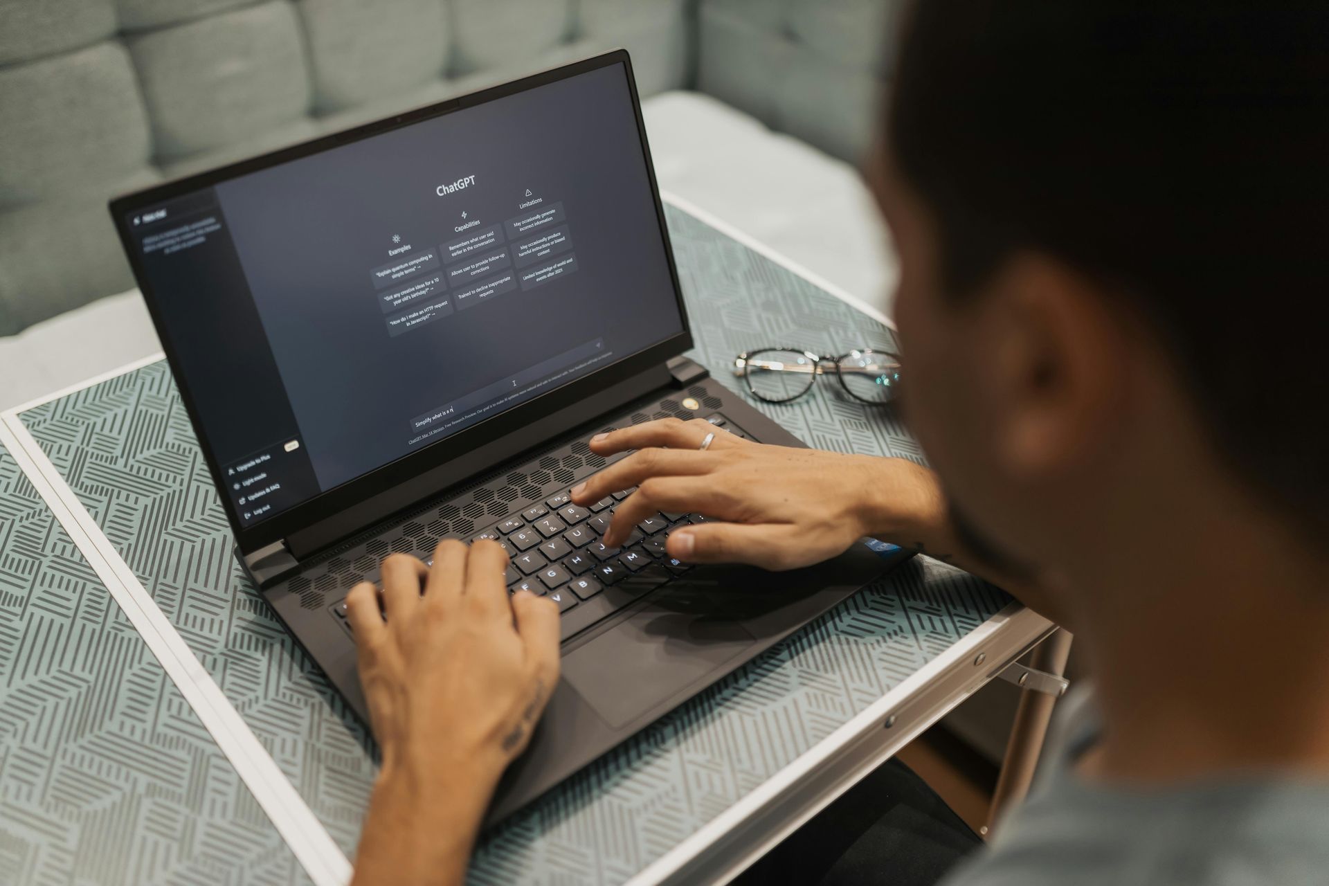 Person typing on a laptop with a dark interface, glasses nearby, on a patterned surface.
