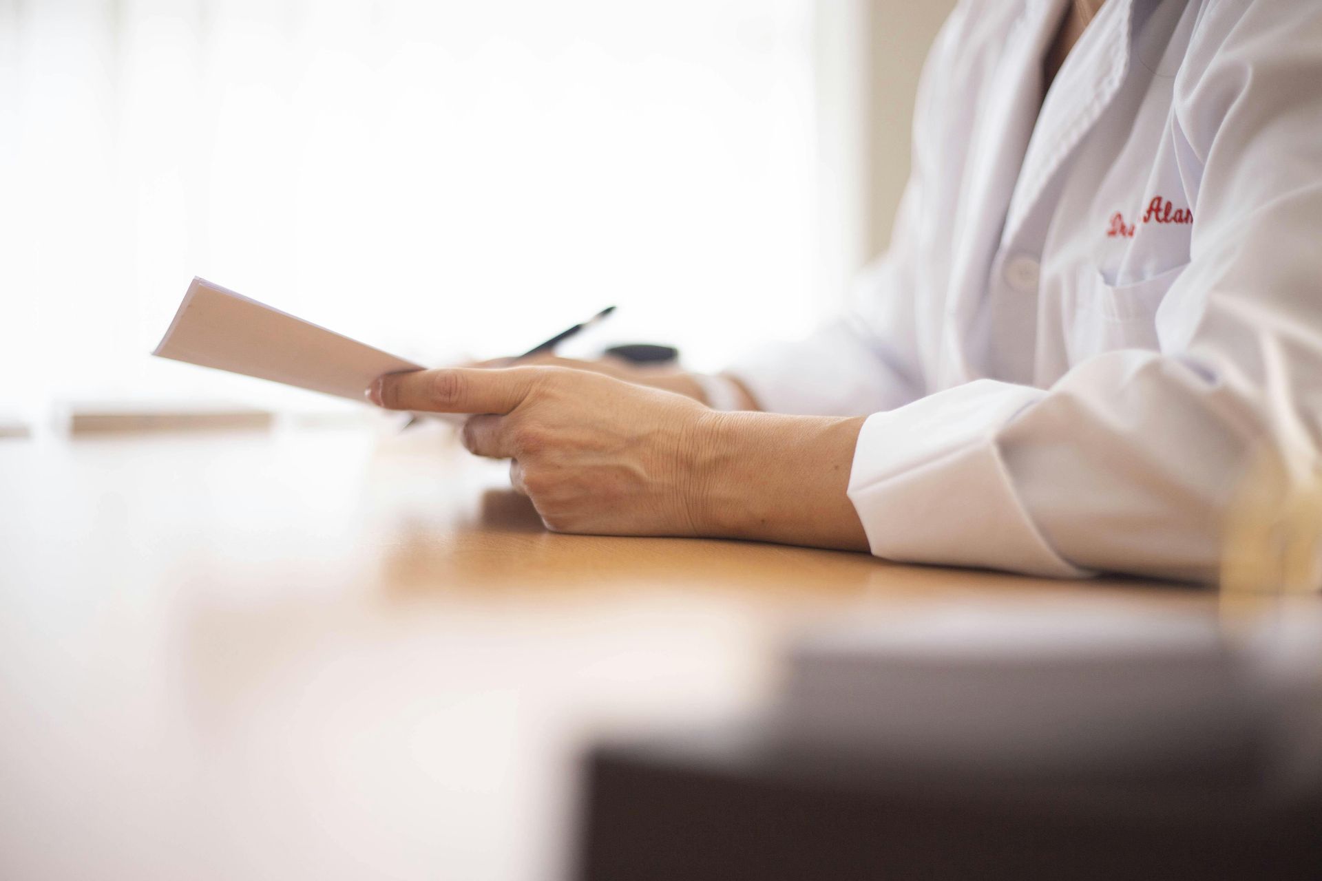 Person in white lab coat holding papers and pen, seated at a desk, likely reviewing documents.