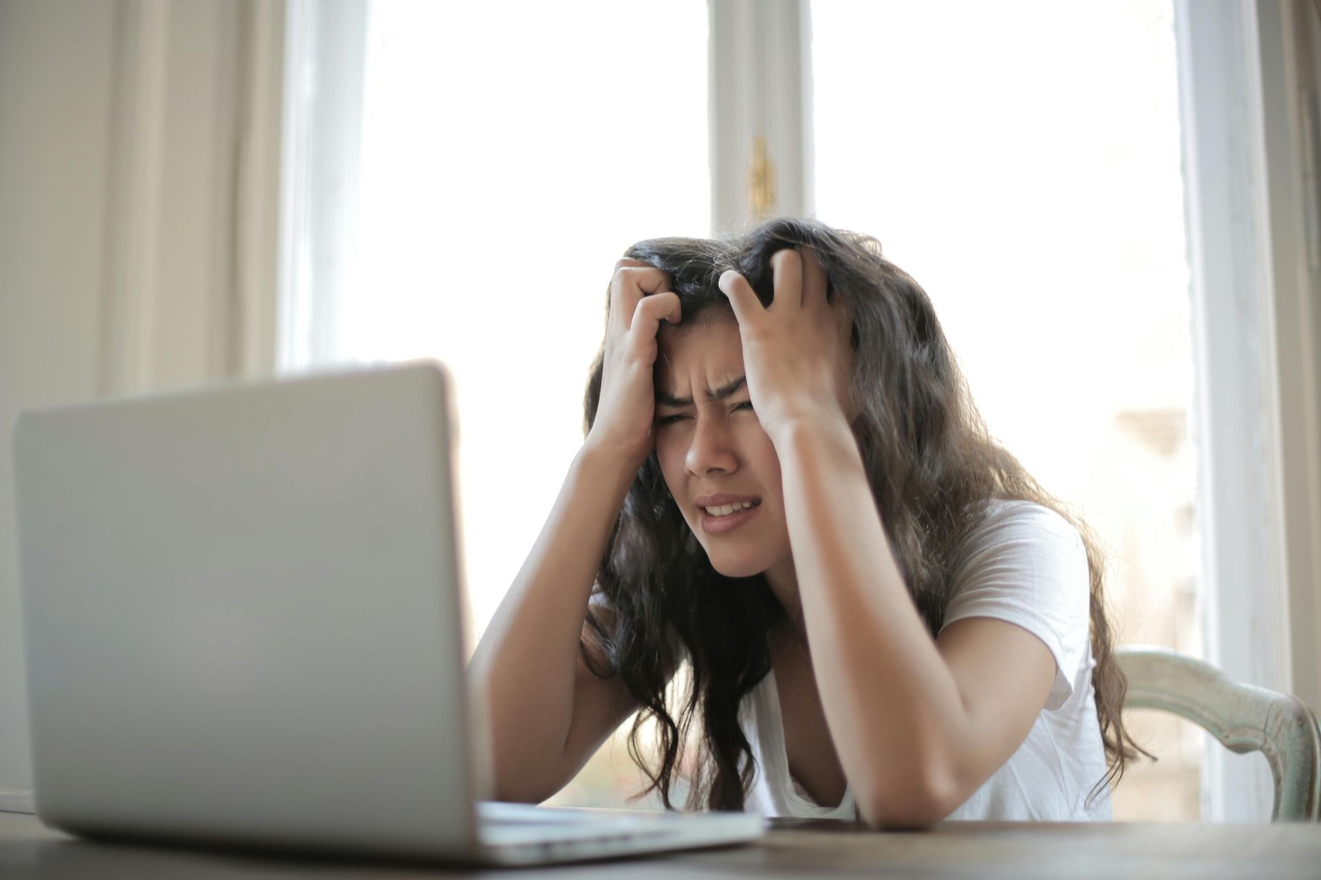 A person with long hair sitting at a table with a laptop, looking distressed while clutching their head.