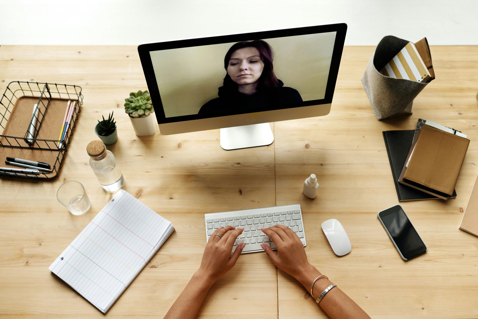 Person typing at computer on video call, wooden desk with notes, phone, water bottle, and potted plant.