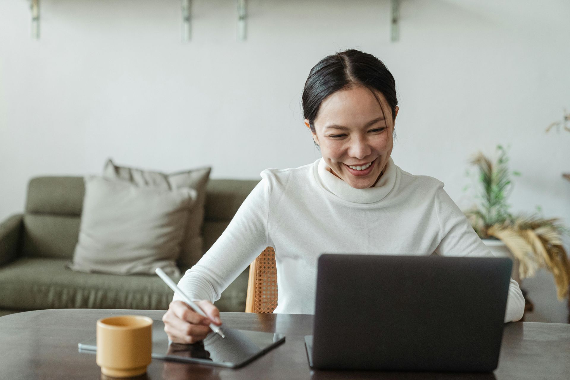Woman smiles while using a laptop and a tablet with a stylus at a table.