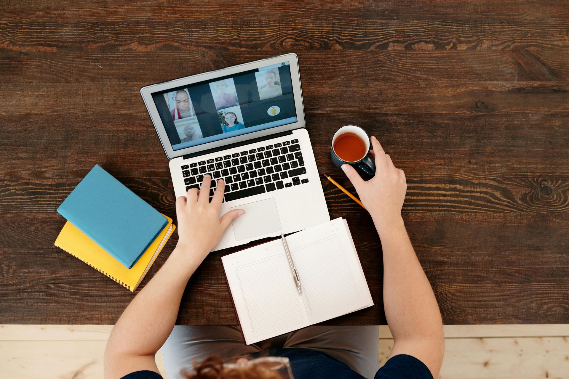 Person working on laptop at a wooden table with a notebook, books, and mug; video call in progress.