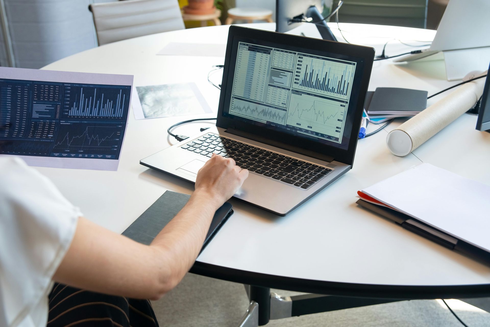 Person at a round table, working on a laptop displaying data, with notes and another screen nearby.