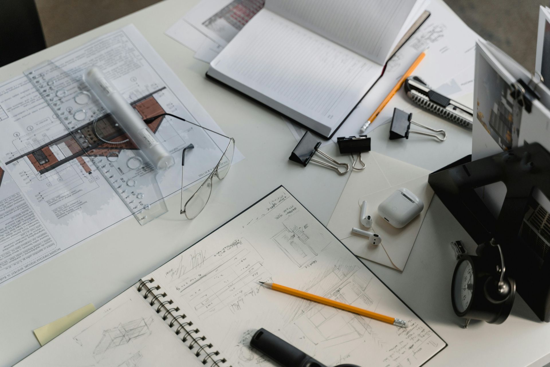A desk with architectural plans, notebooks, pencils, and an alarm clock, suggesting work in progress.