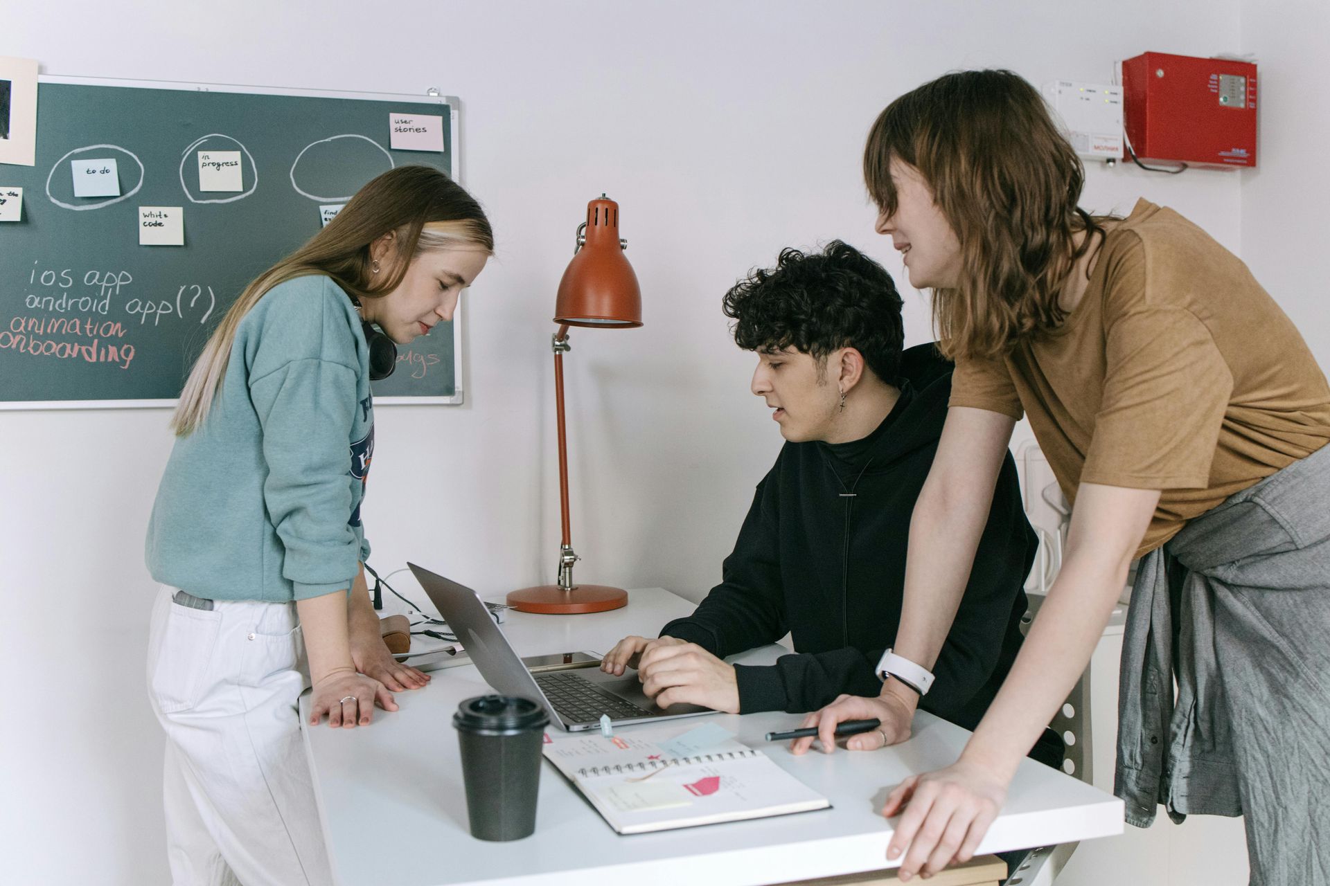 Three people collaborating around a laptop at a desk with a coffee cup.