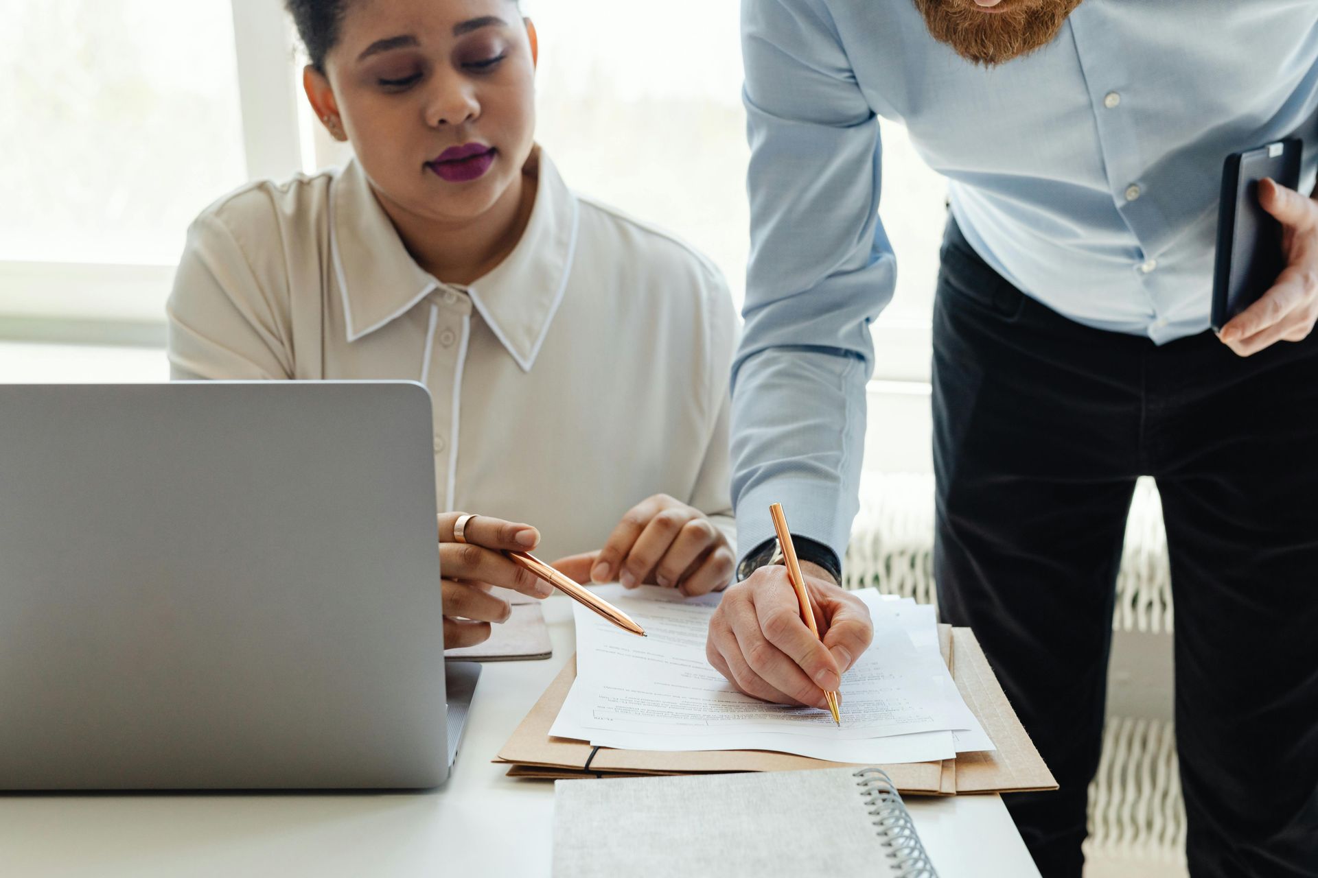 A person is signing a document while another person points at the document next to a laptop.