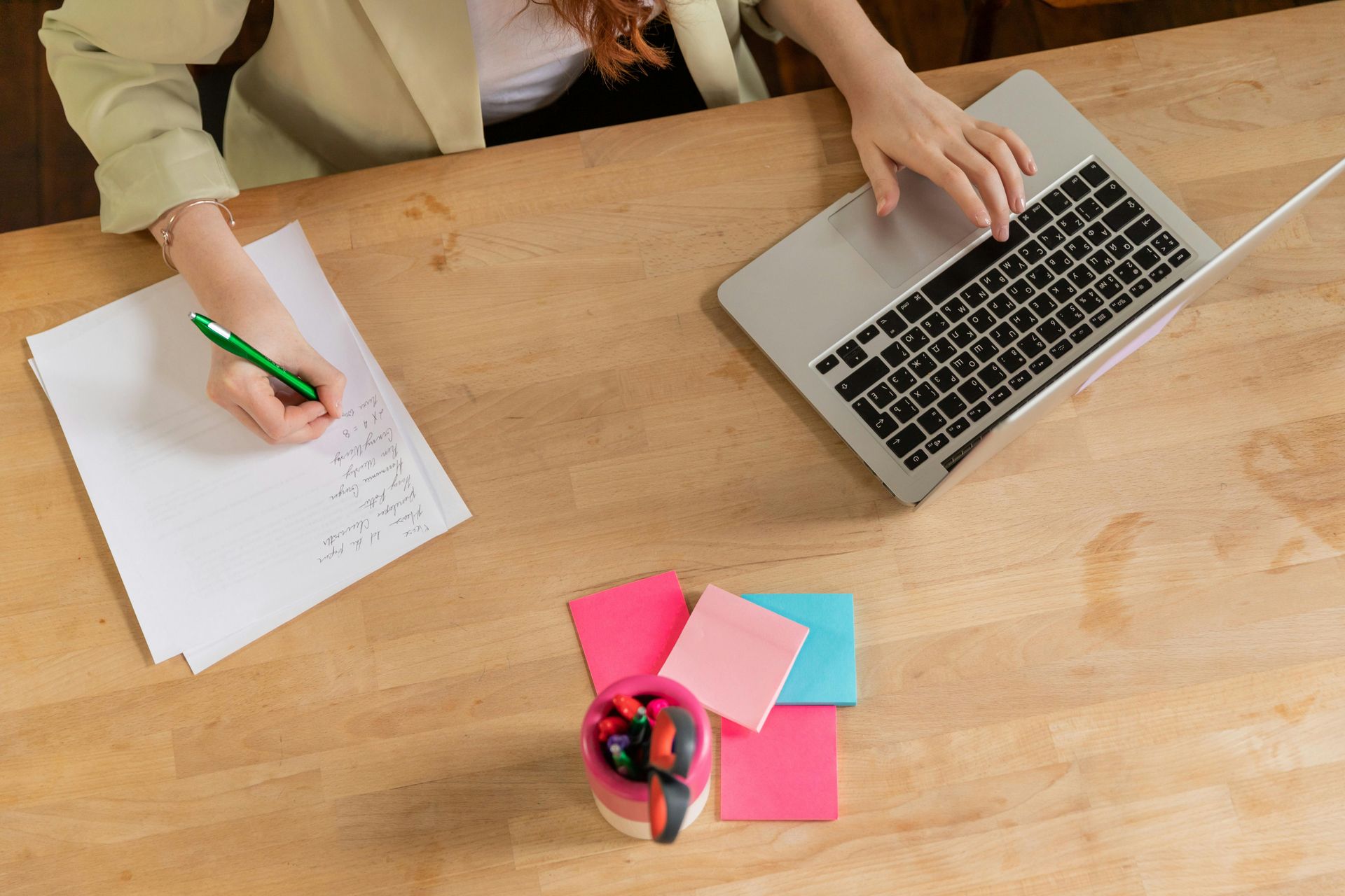 Person working at a wooden desk, writing on paper with a pen and typing on a laptop. Sticky notes and pens are nearby.