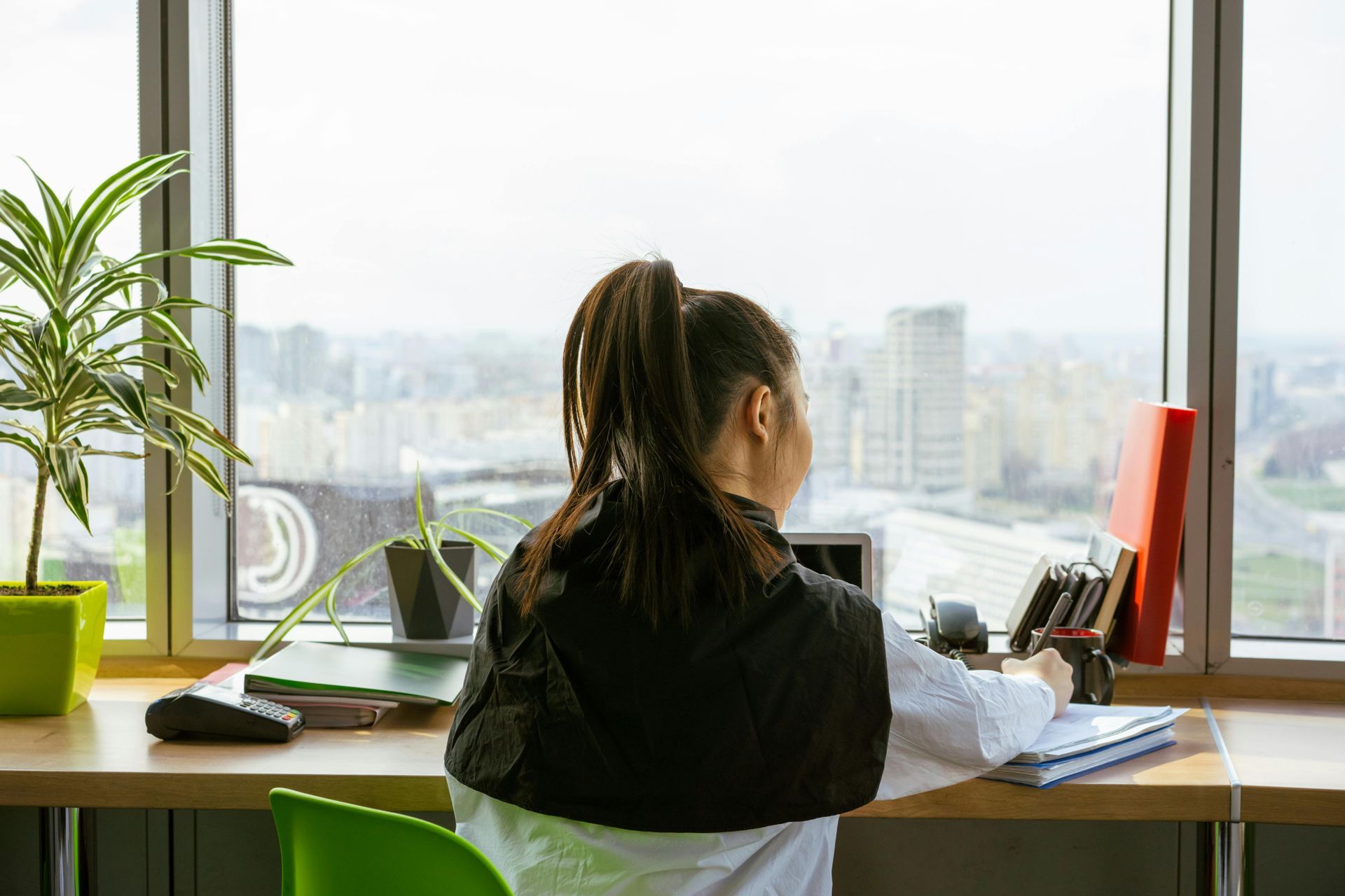 Woman at desk, working near a window with city view. Green plant and red monitor present.