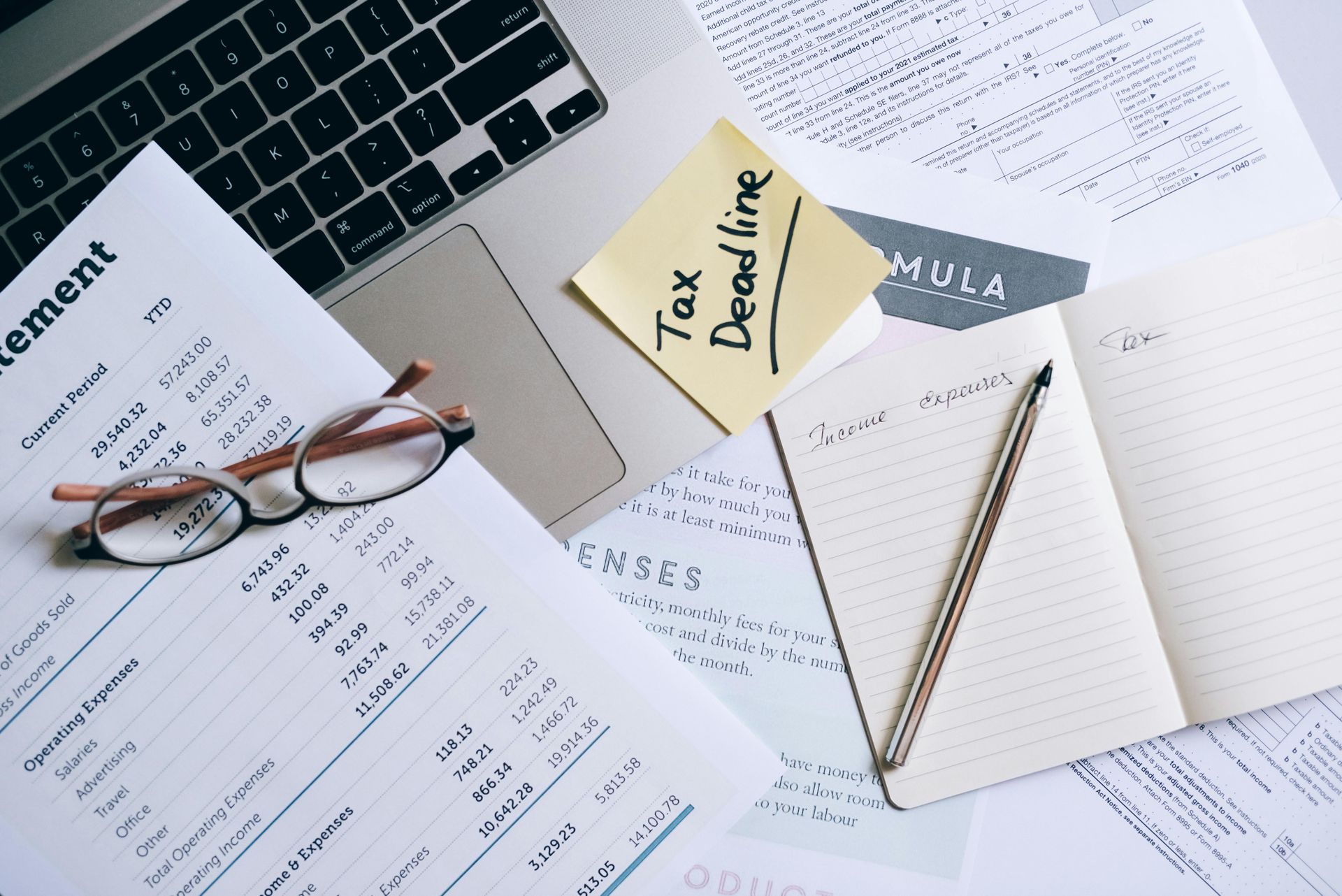 A laptop, a document with a tax deadline sticky note, eyeglasses, and an open notebook with a pen on a desk.