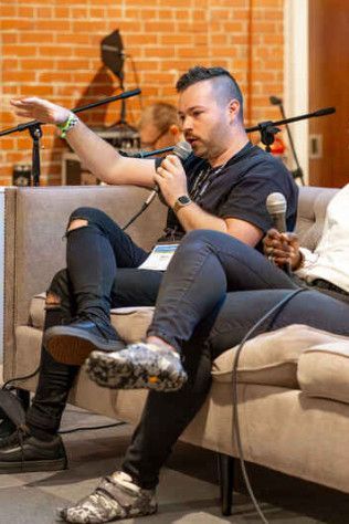 A person sits on a sofa on stage, holding a microphone and gesturing while speaking at a panel discussion.