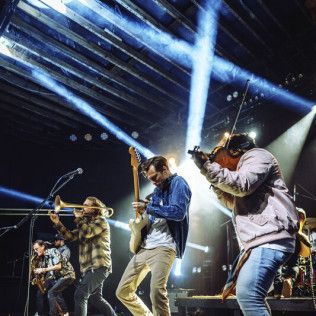A live band performs on stage under bright spotlights, featuring a violinist, a guitarist, and a brass player.
