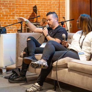 Two people sit on a sofa at an indoor event, talking into microphones in front of a brick wall.