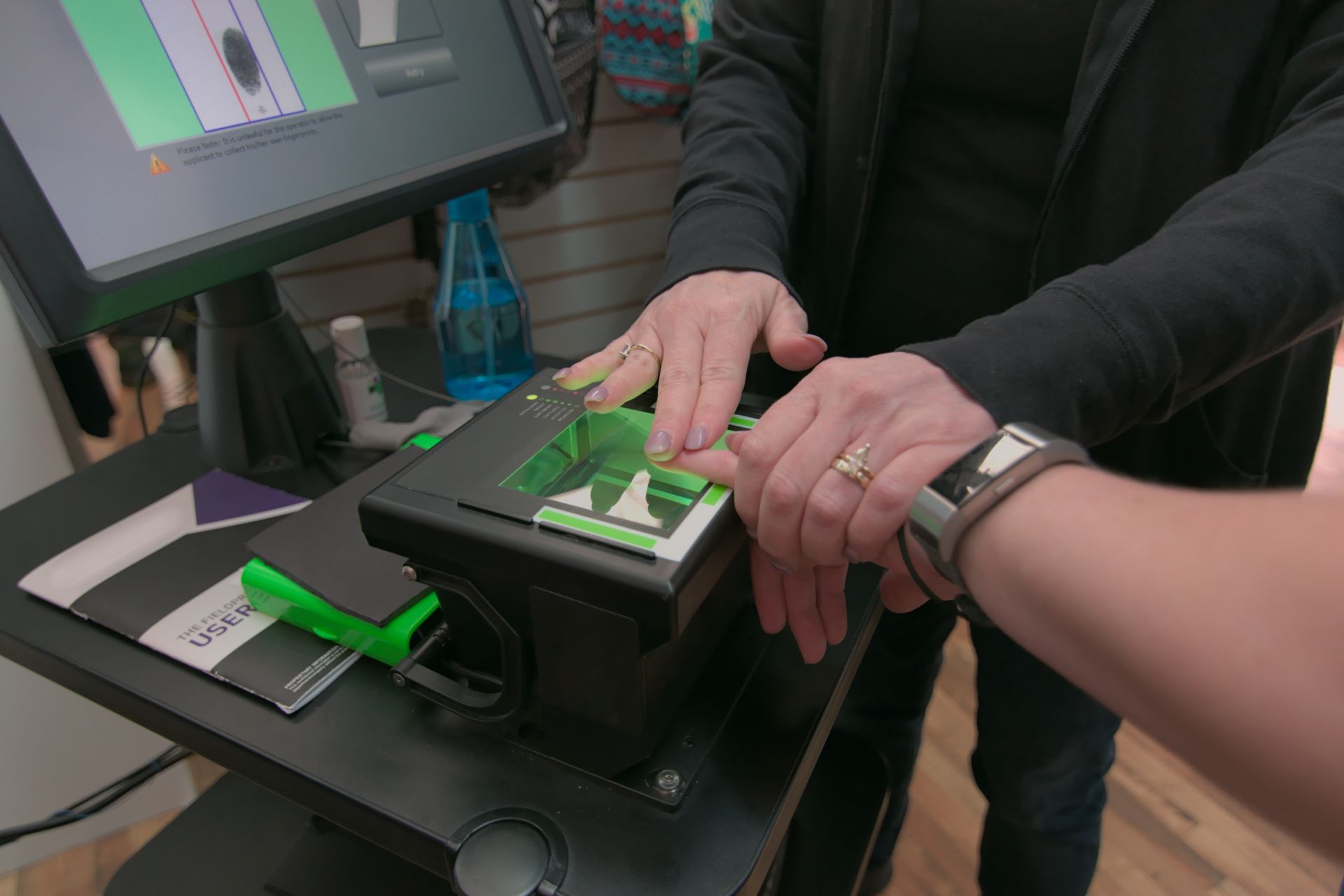 Person scanning a fingerprint on a black device. A screen and other items are nearby.