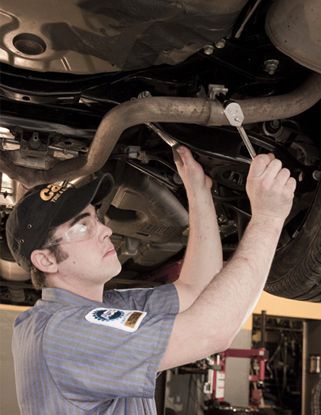 Mechanic working under a car, using a wrench on exhaust system. Wearing glasses, cap, and grey uniform.