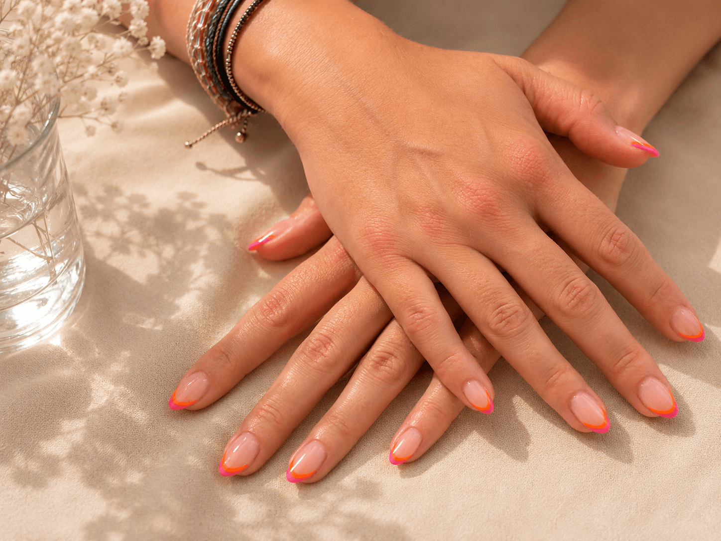 Hands with coral-pink nails resting on a satin surface beside a glass candle