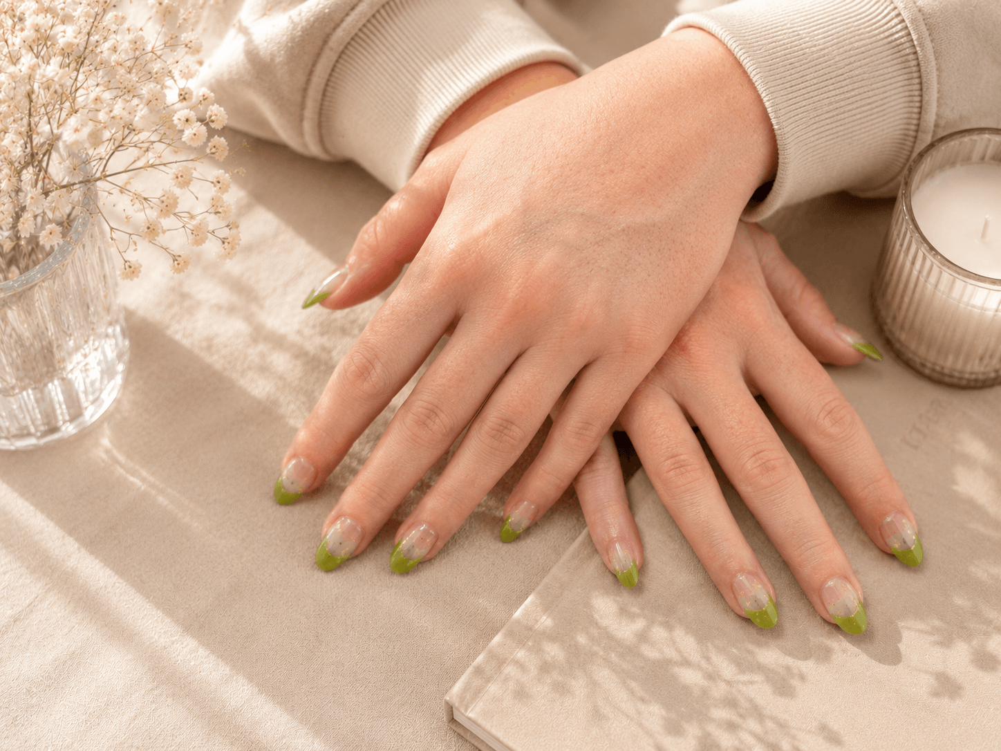Hands with green nails resting on a beige fabric surface near a small cream candle.