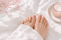 Bare feet resting on white bedding beside a pink candle and delicate flowers
