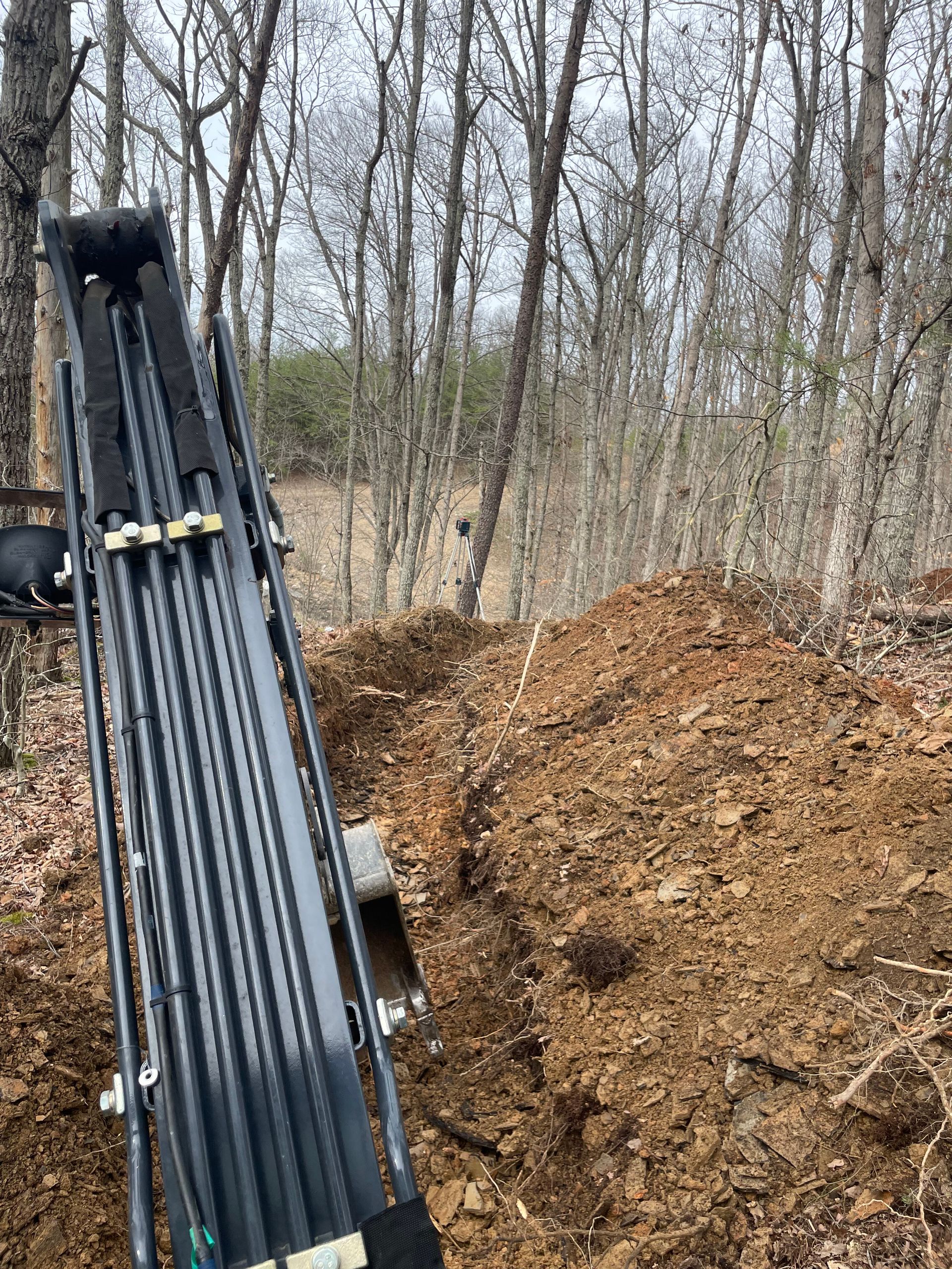Black machinery boom beside a large dirt pile in a leafless wooded area