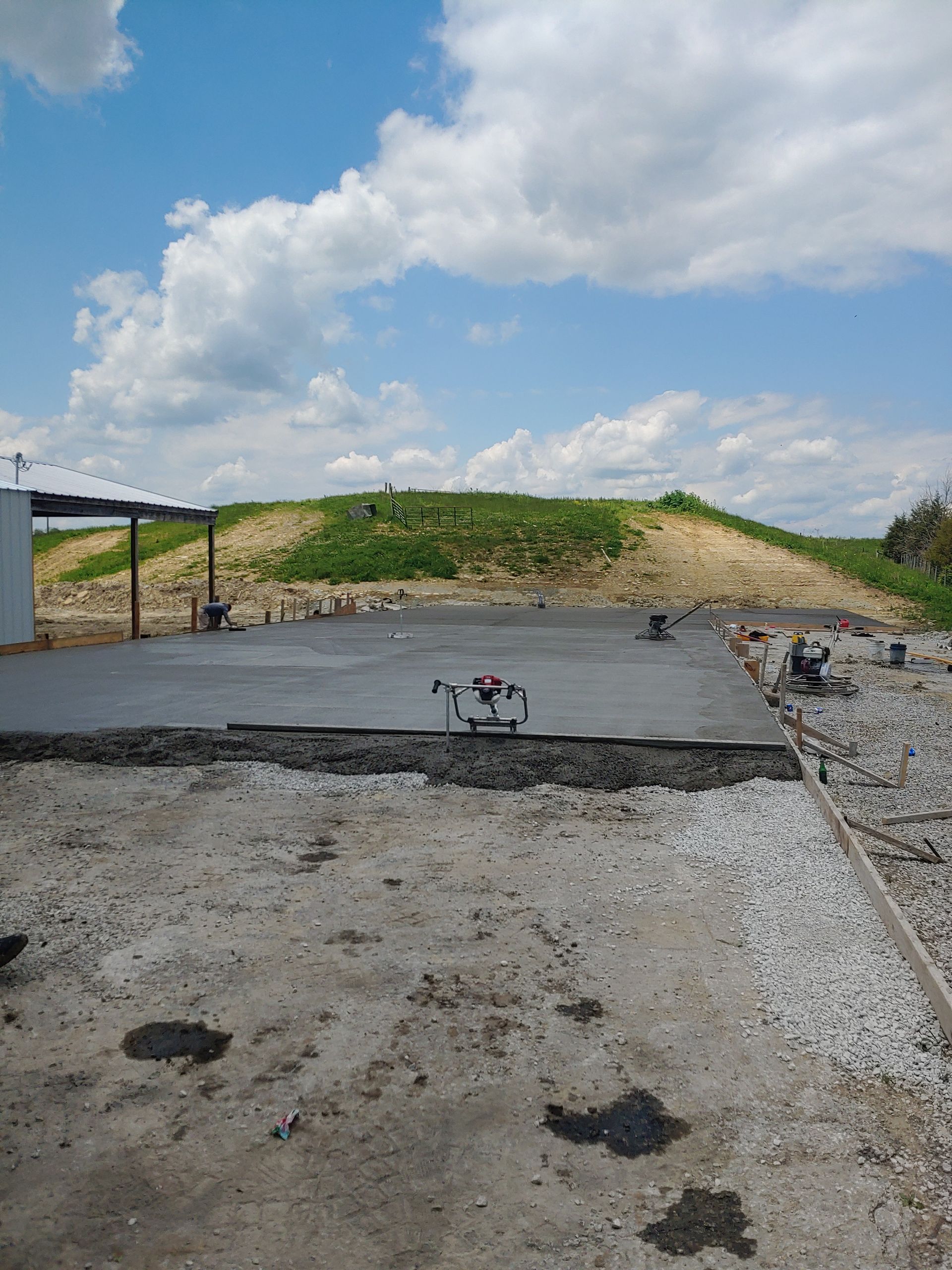 Gravel lot with a black paved strip, small dog in the center, and a grassy mound under a cloudy blue sky