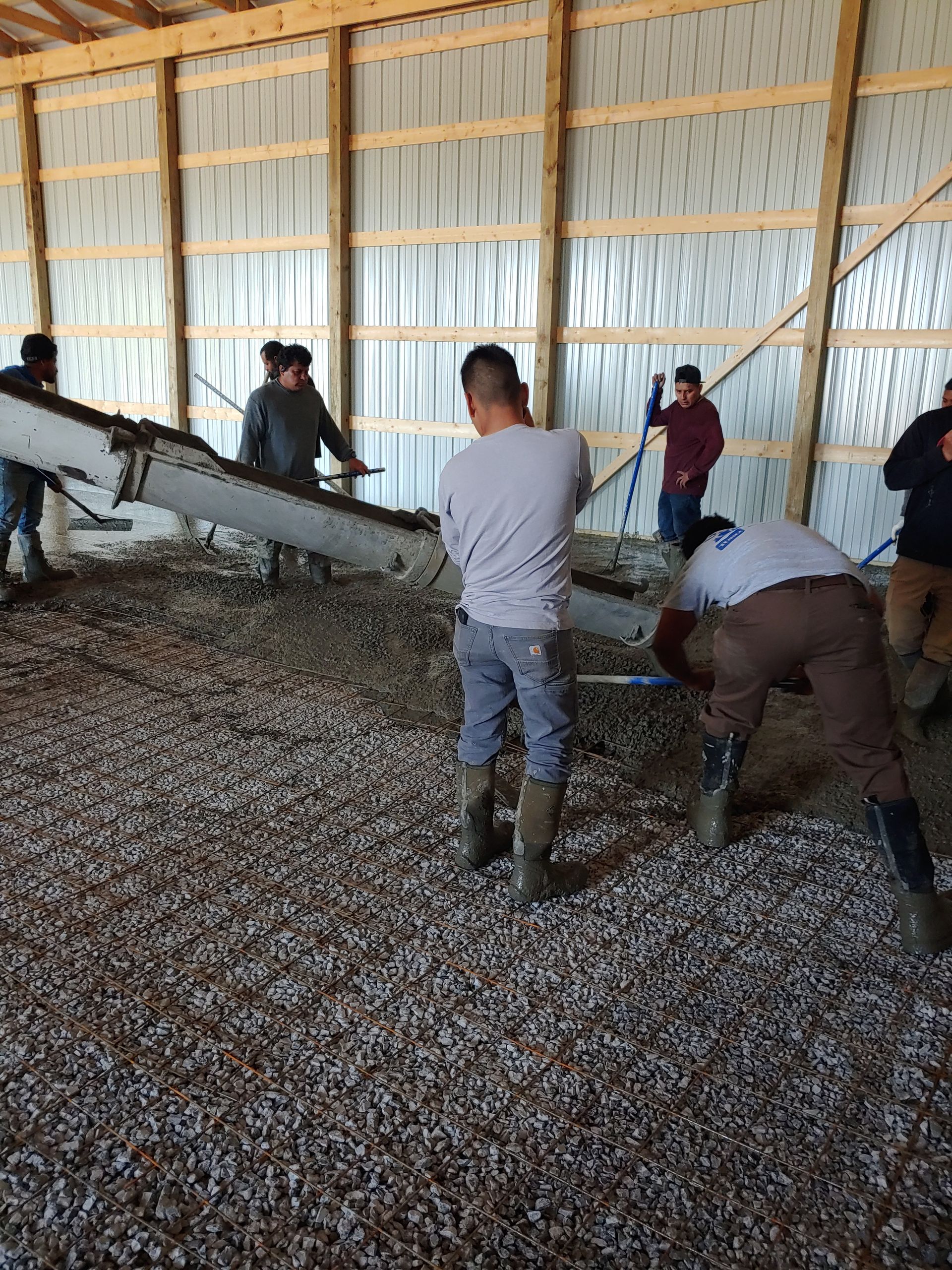 Workers leveling gravel inside a large wooden shed with a conveyor belt unloading material