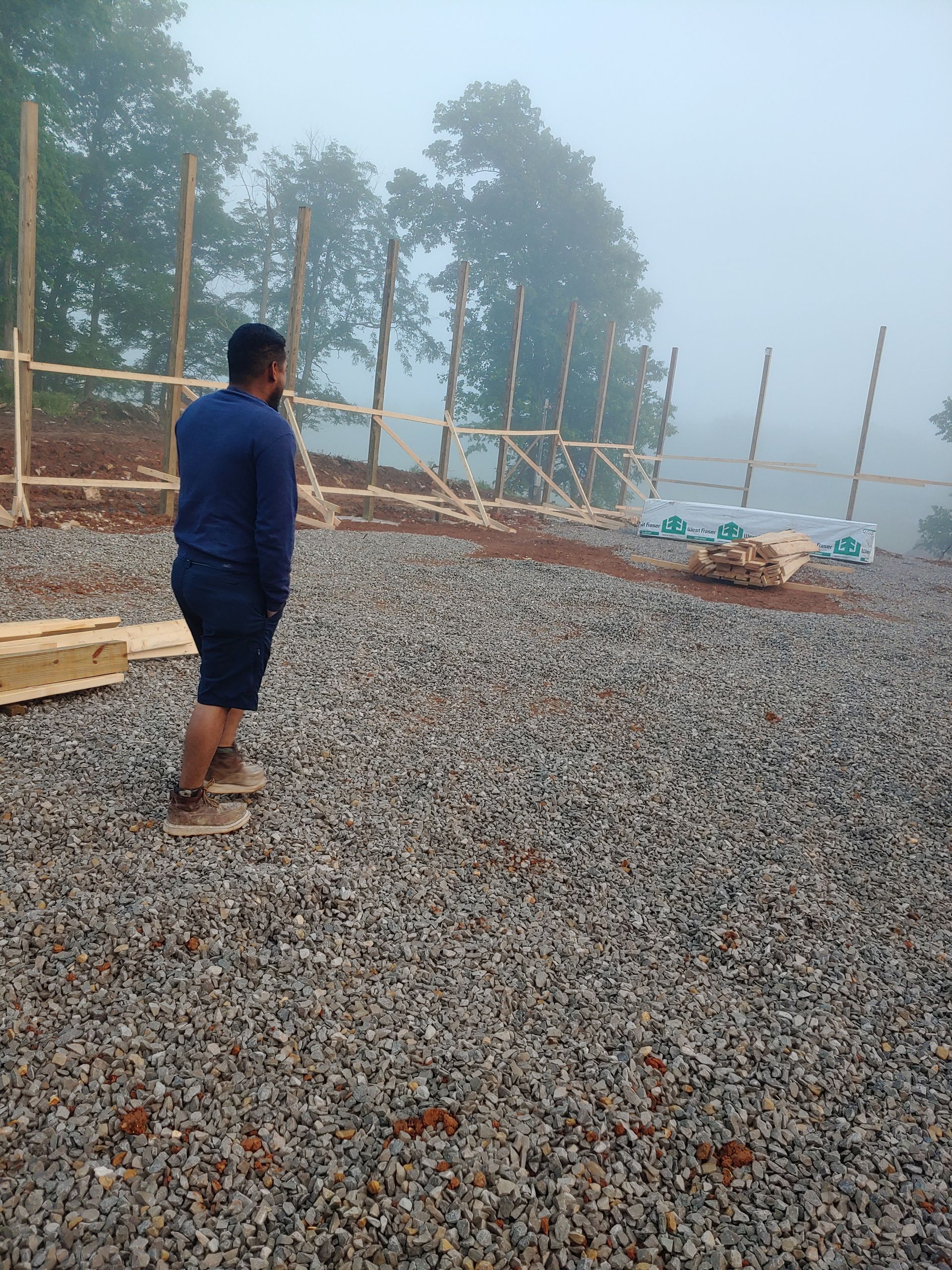 Person standing on a gravel foundation at a misty construction site with wooden posts and trees in the background