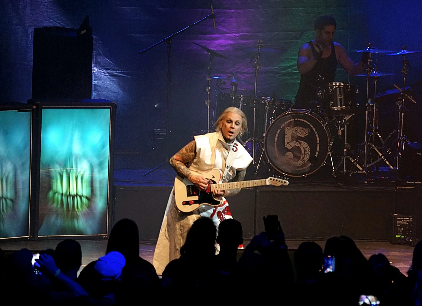 Singer playing guitar on a dimly lit stage with blue and purple lights, drummer in the background.