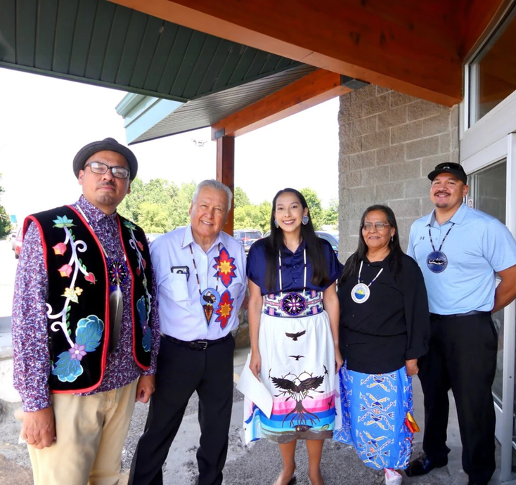 Five people posing under a covered walkway, wearing traditional and formal clothing and smiling.