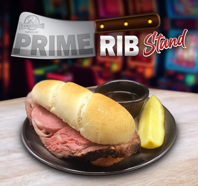 Prime Rib sandwich on a black plate with au jus and pickle, under a “PRIME RIB Stand” sign.