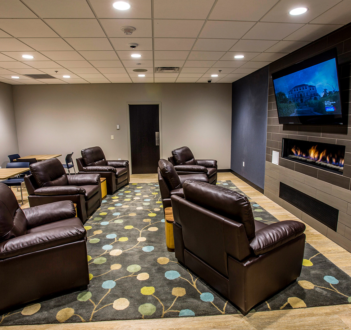 Modern lounge with brown leather chairs, patterned carpet, and a wall-mounted TV above a fireplace.