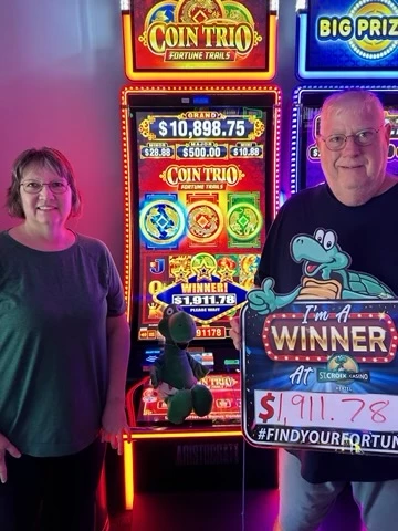 Two people standing beside a bright Coin Trio casino machine, one holding a “I’m a winner” sign