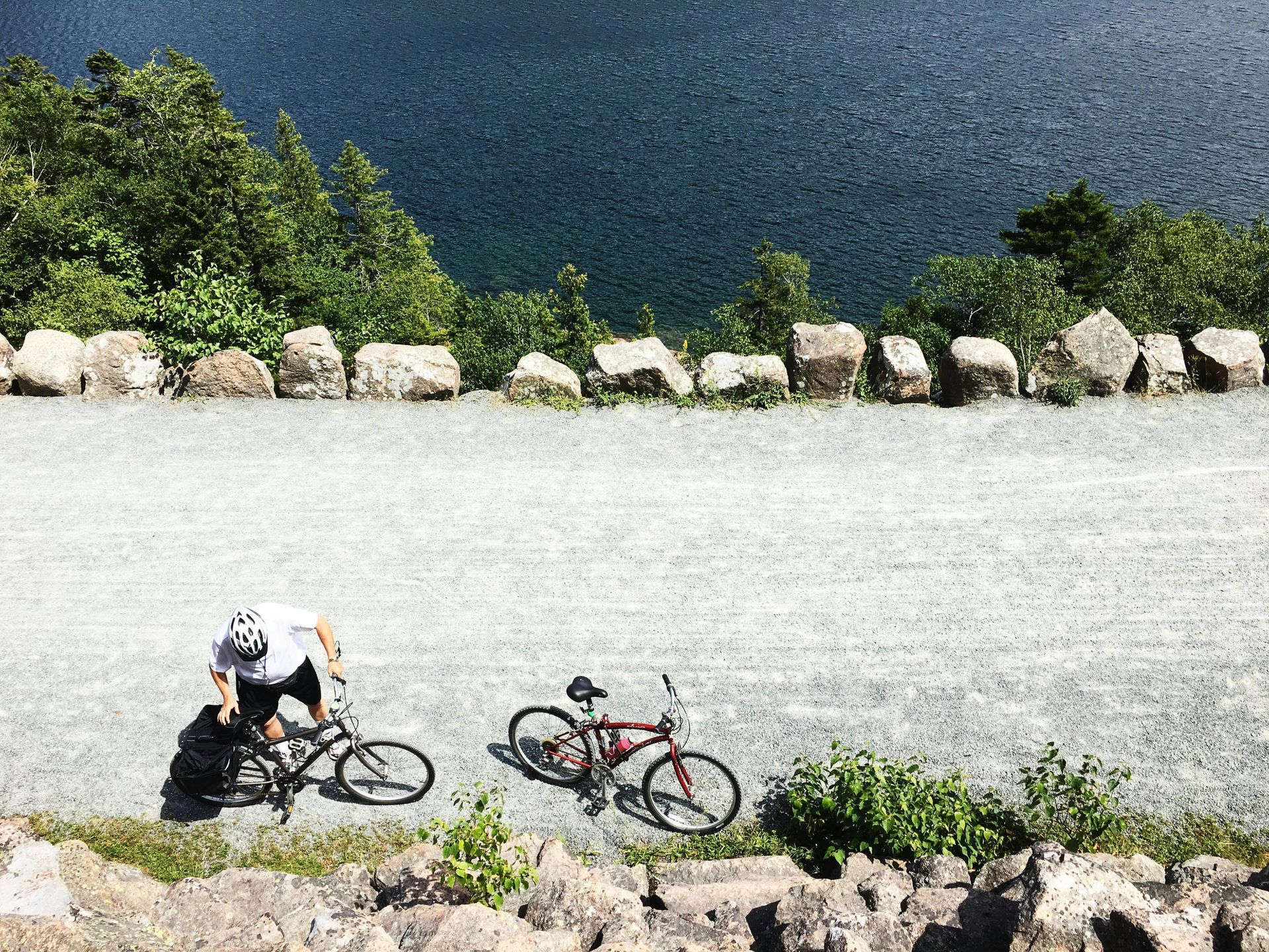 Two cyclists with road bikes on a gravel path beside blue water and rocks