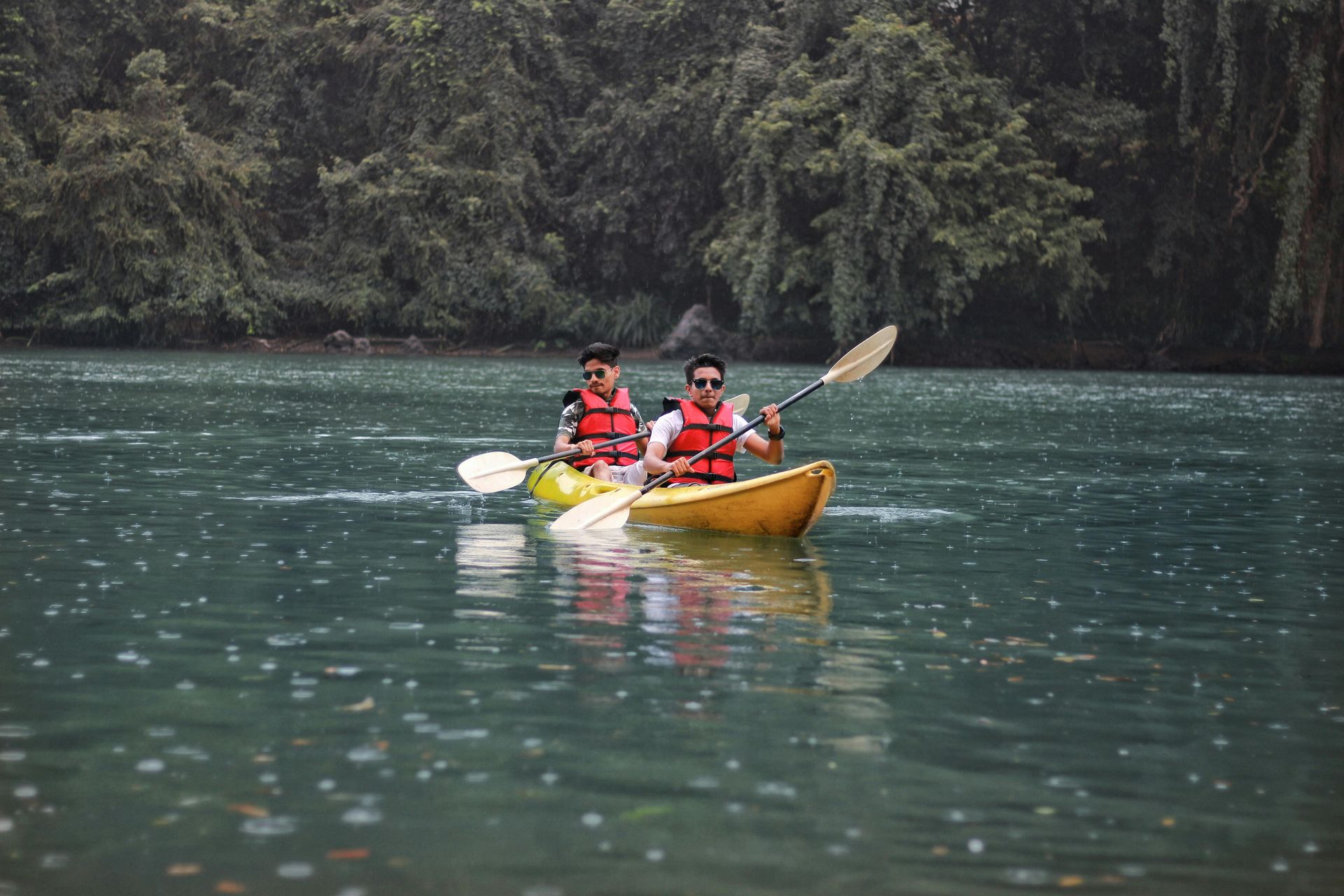 Two people in red life jackets kayaking on a calm green river, surrounded by dark forest.