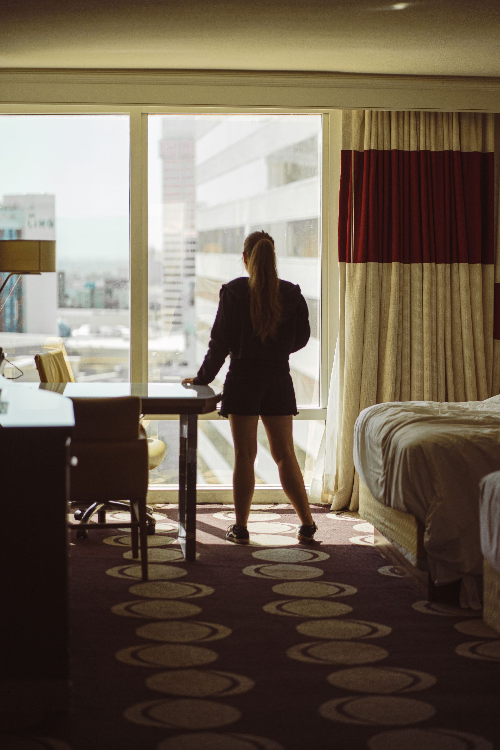Silhouette standing by a hotel room window, looking out over the city, with a bed and desk nearby