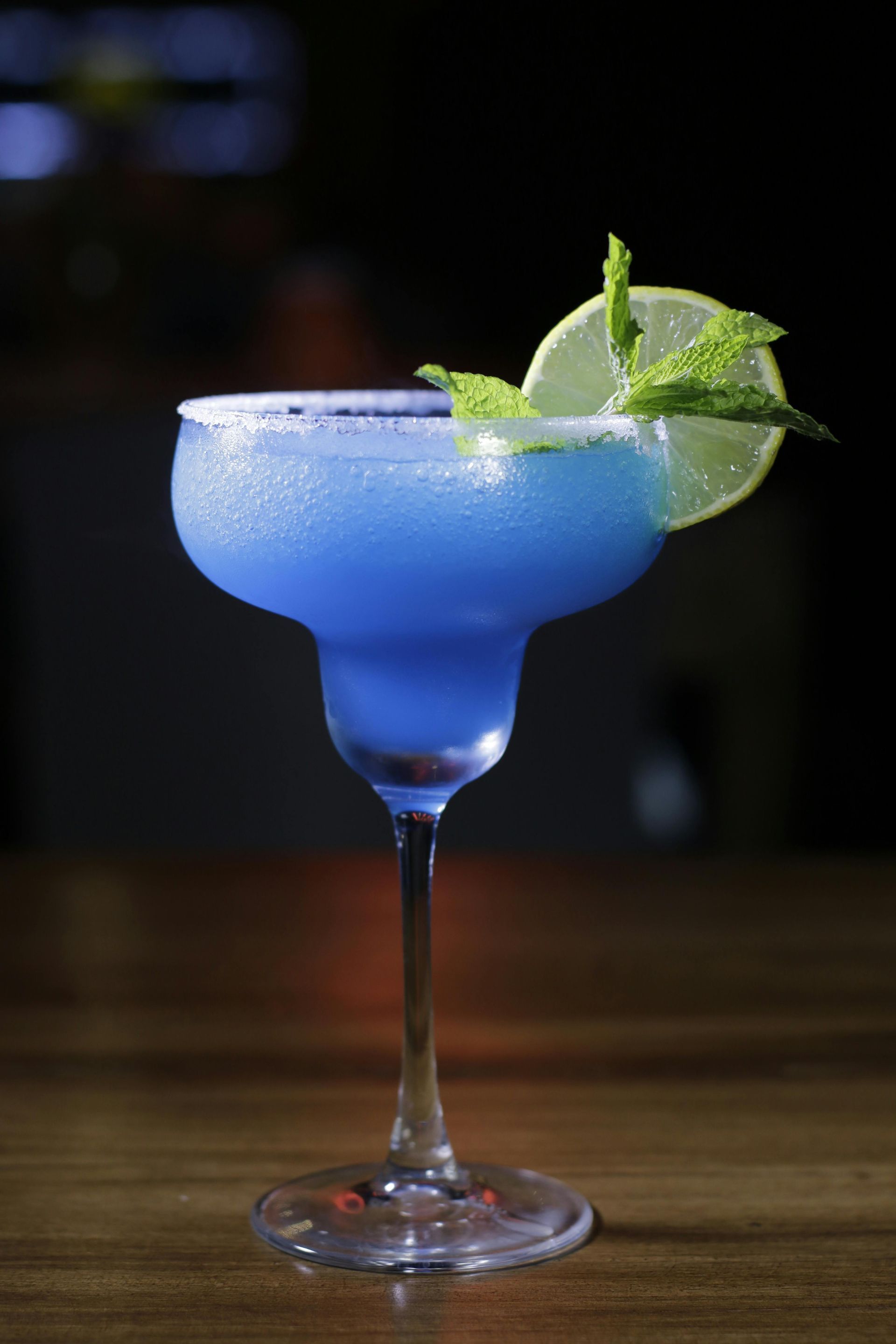 Blue cocktail in a stemmed glass with mint and lime garnish on a wooden table