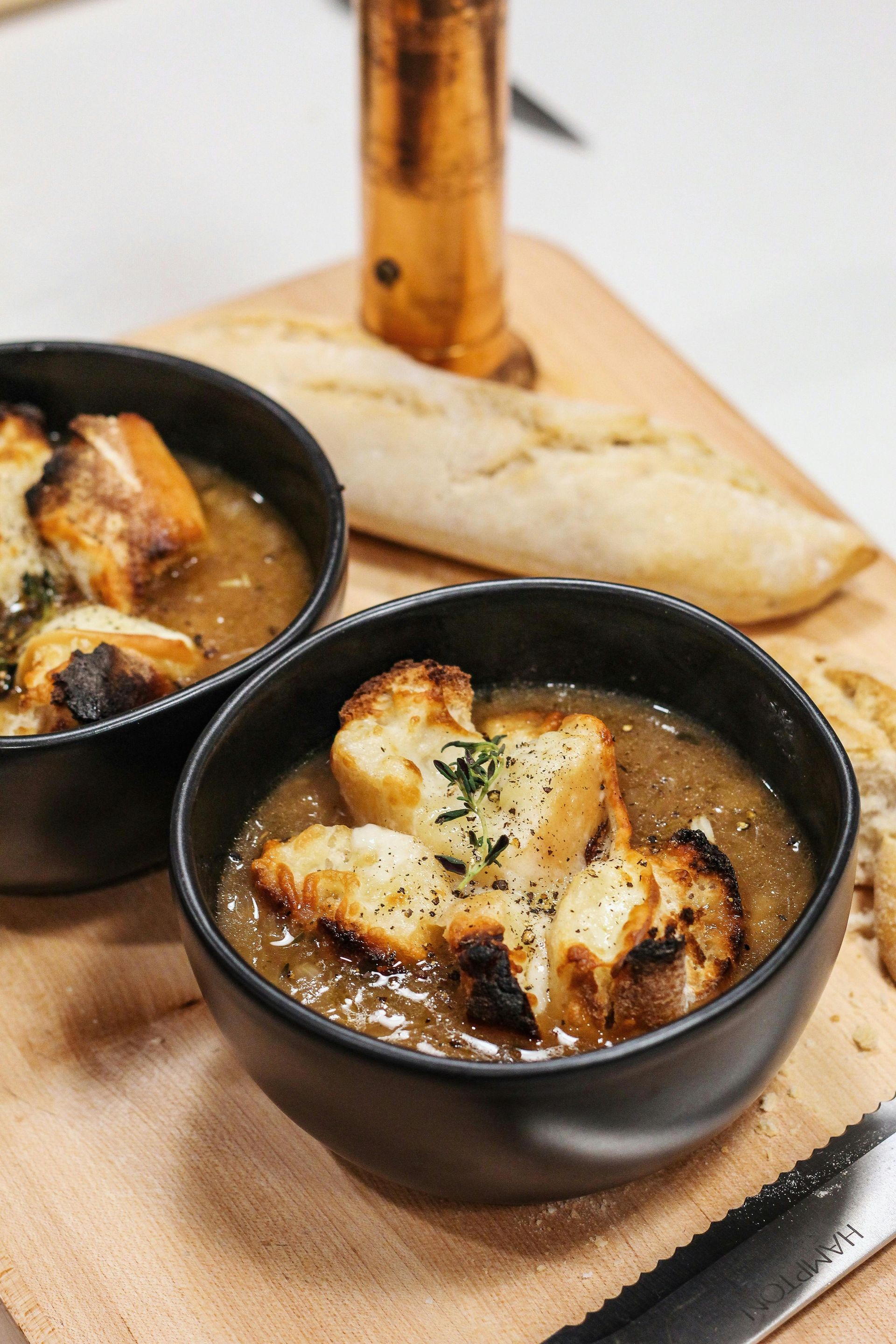 Two bowls of baked soup topped with melted cheese on a wooden board, with bread in the background