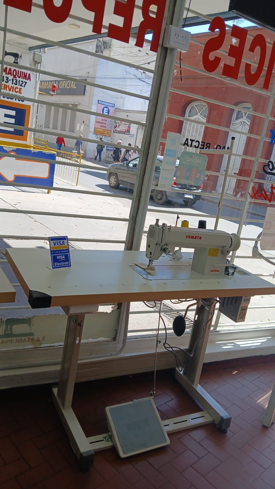 Máquina de coser blanca sobre una mesa junto a un luminoso escaparate con letras rojas en el exterior.