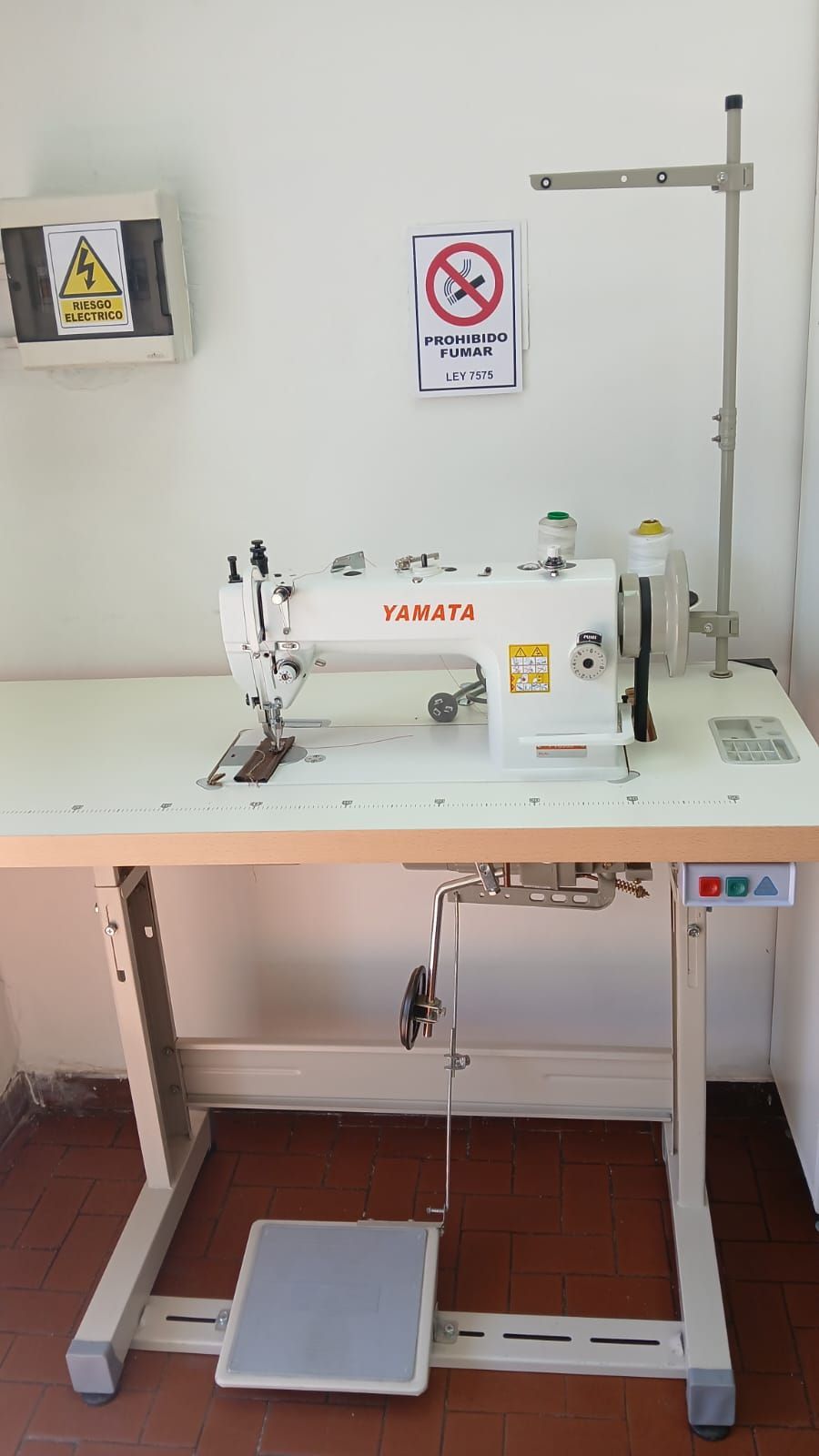 Máquina de coser montada sobre una mesa motorizada ajustable con pedal y barra de soporte superior.