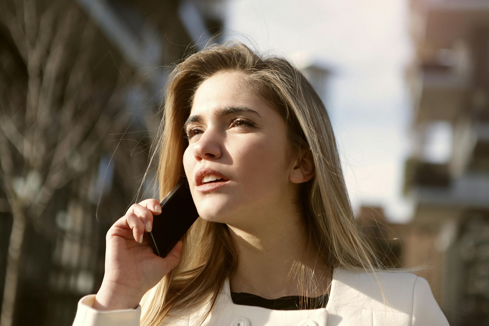 A person with long blonde hair wearing a light jacket talks on a smartphone while outdoors in a city setting.
