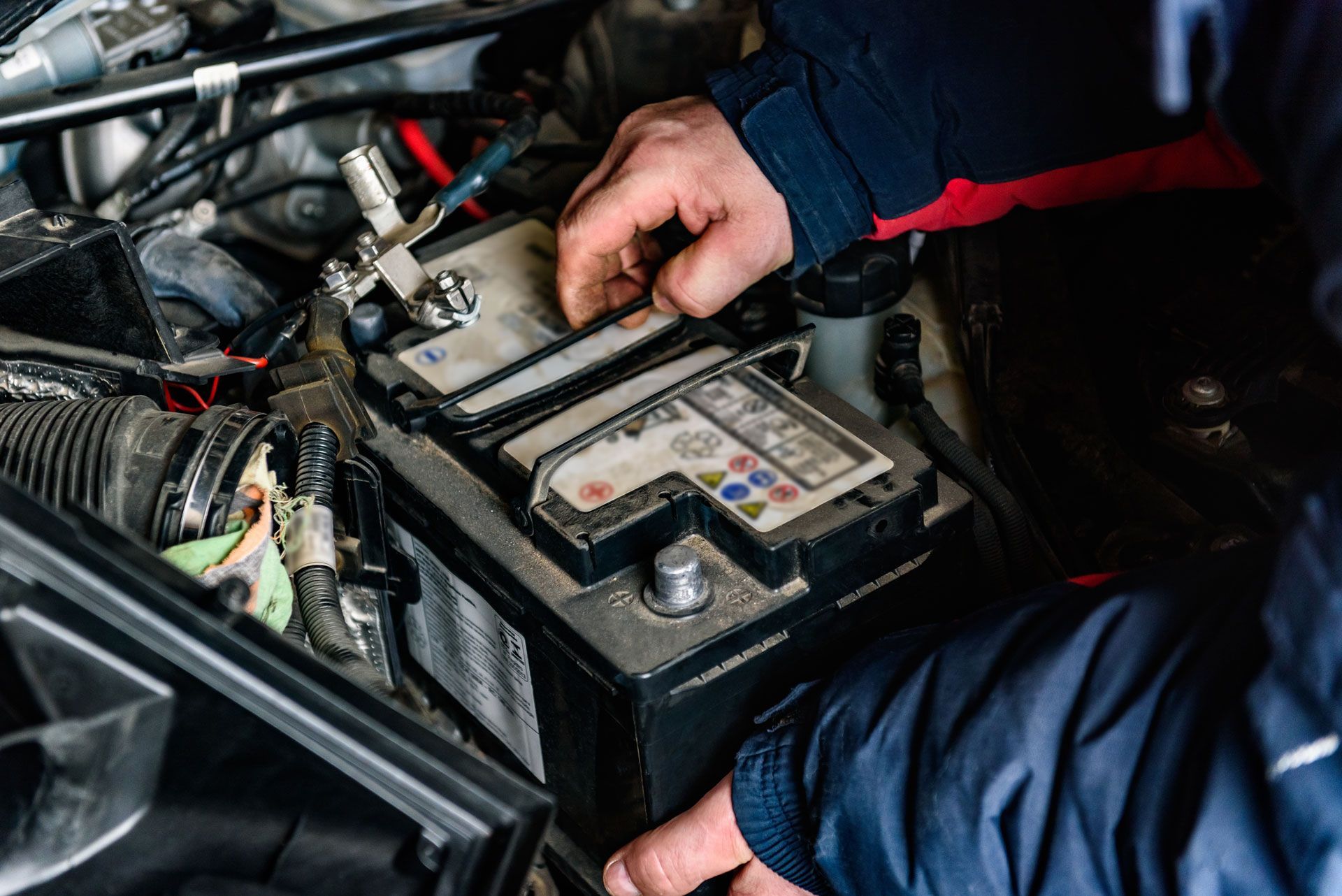 Mechanic testing a car battery under the hood with wires and tools visible