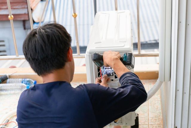 Man using a power drill to work on white electrical equipment, outdoors on a balcony.