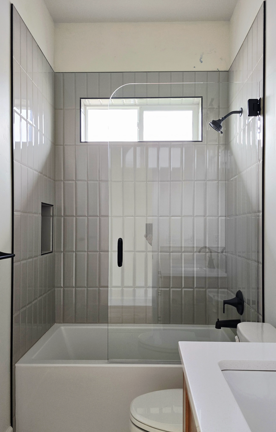 Bathroom with light gray tile, tub, glass shower door, black fixtures, and a small window.
