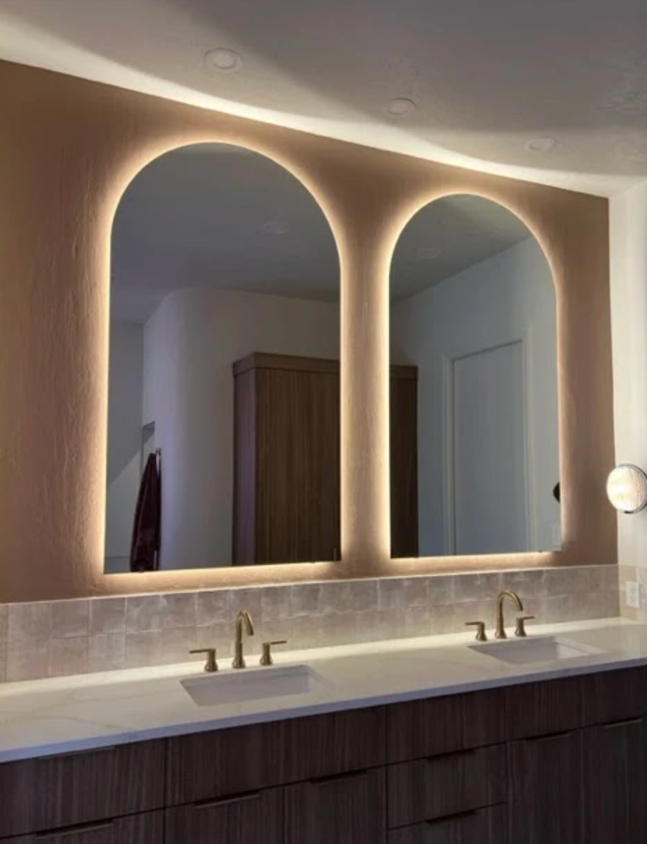 Two arched, backlit mirrors mounted over a double vanity with wood cabinets and white tile backsplash.