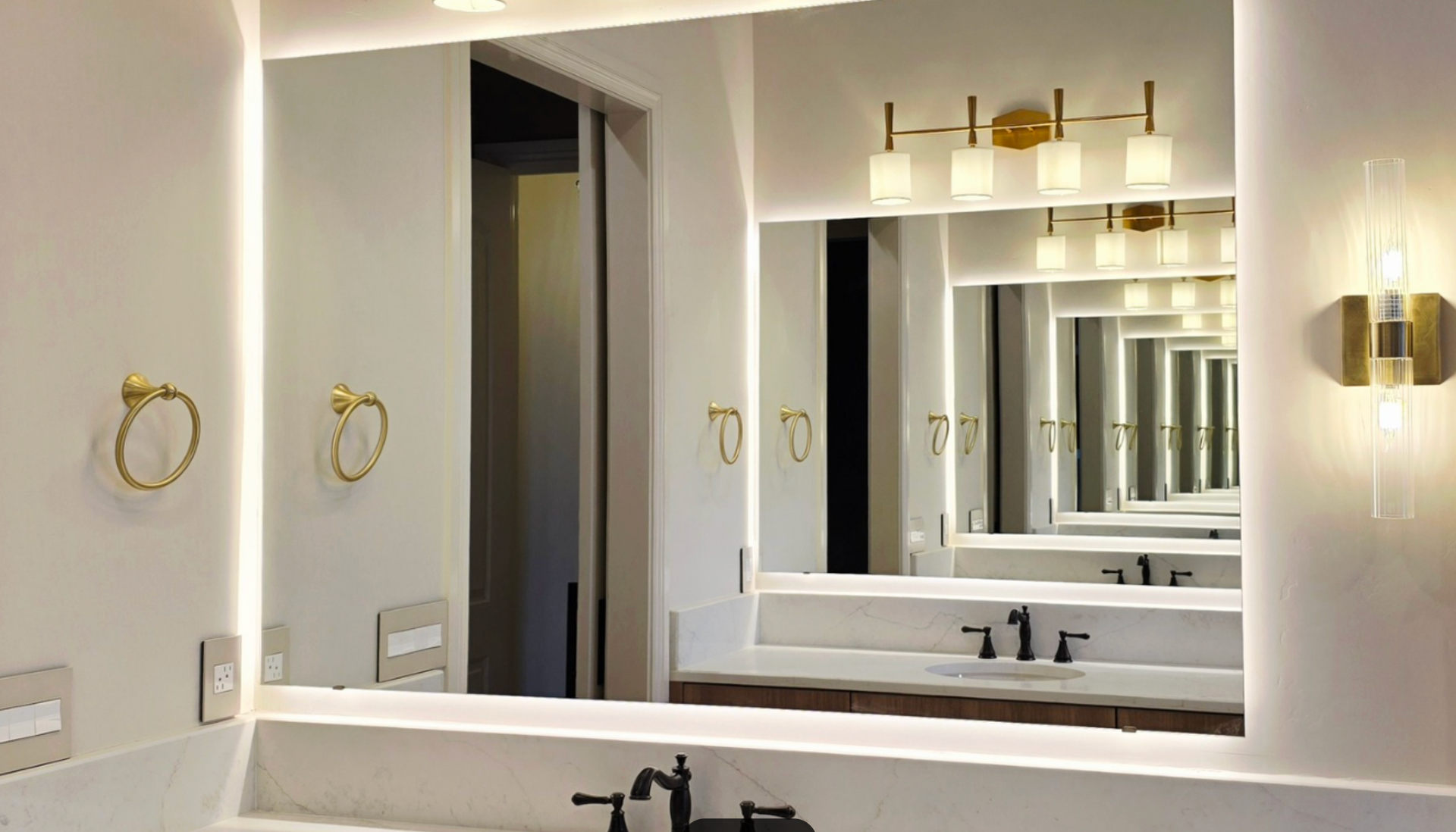 A modern bathroom vanity with a large, LED-backlit mirror, brass hardware, and wall sconces against neutral-toned walls.