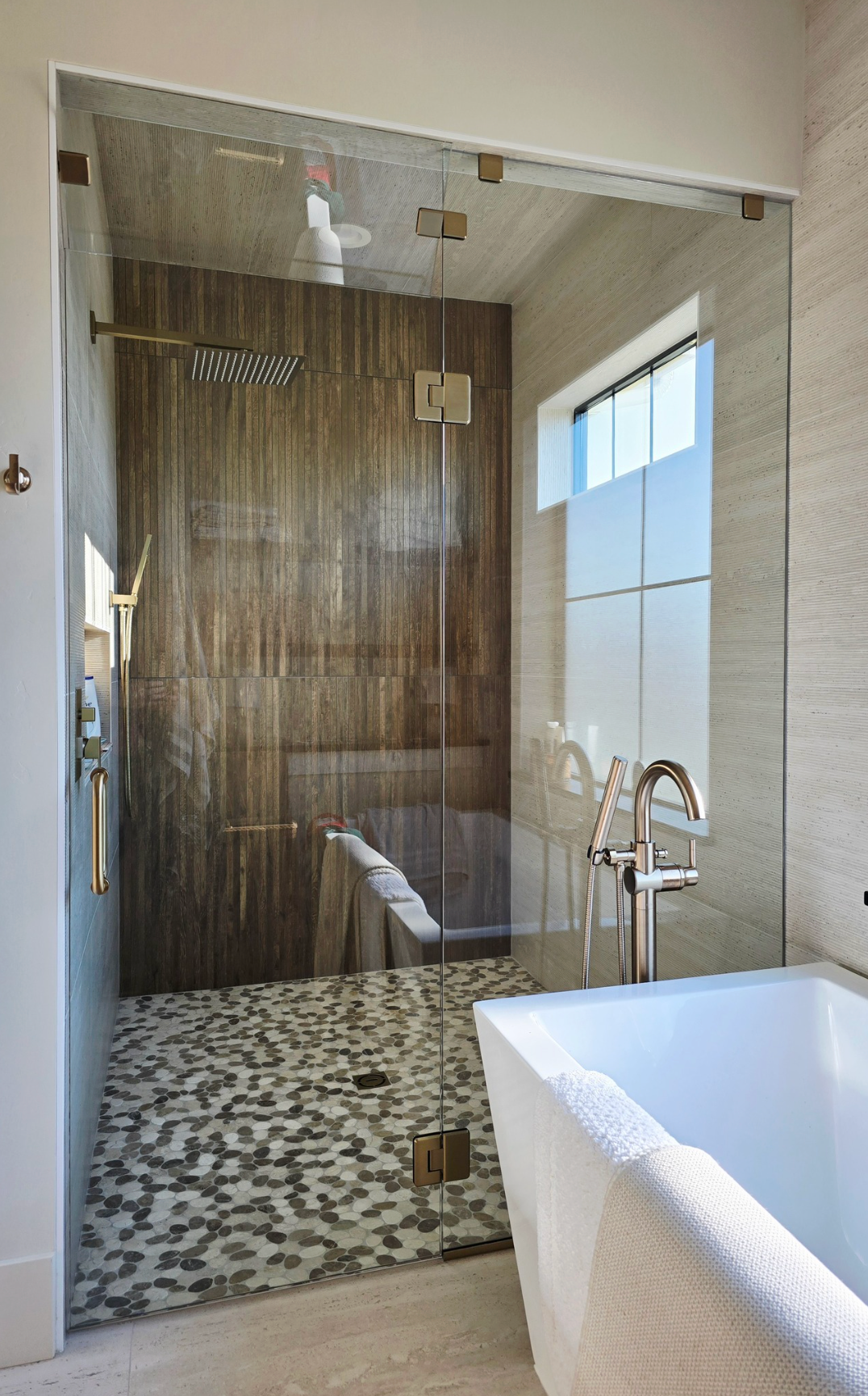 A glass-enclosed shower with dark wood-look tile, a pebble floor, and a white freestanding tub in a bright bathroom.